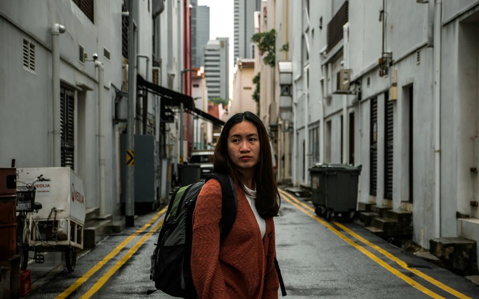 A woman with a backpack pauses in a quiet alley between city buildings, looking back over her shoulder