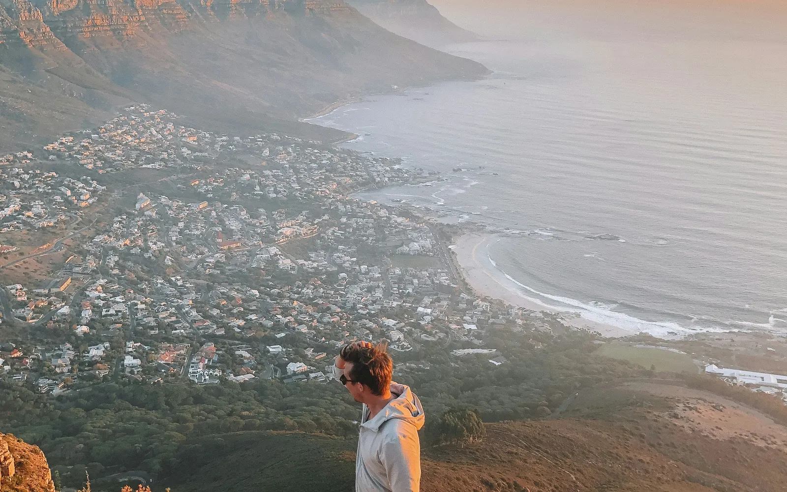 A hiker looking out over a coastal city and ocean from a mountain summit at sunset